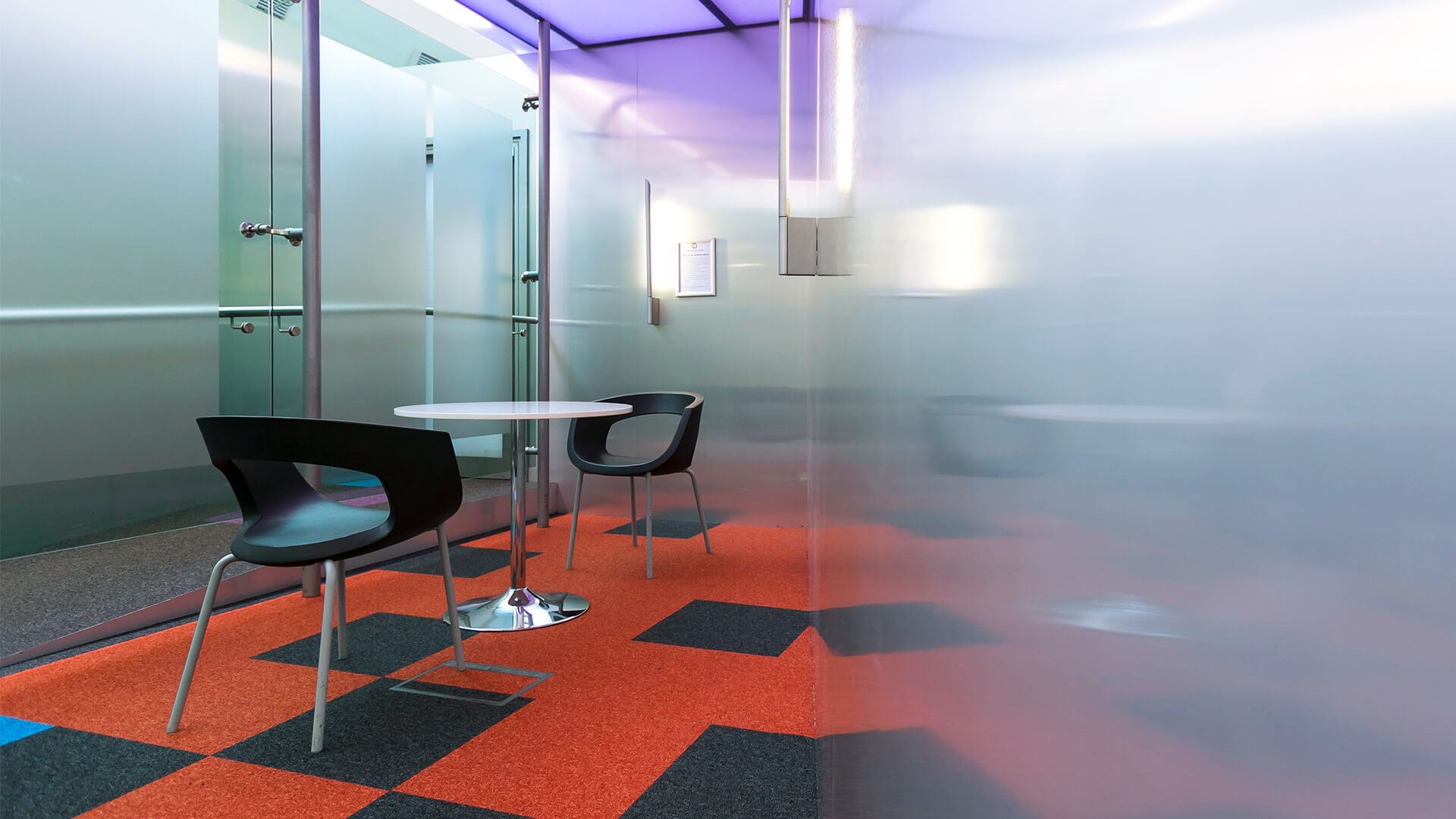 Glass and brushed steel walls enclose a bright breakout meeting space with a table and chairs
