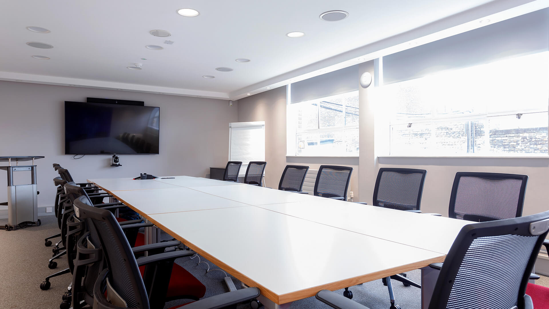 A large screen is mounted at the head of the table in a light-filled meeting space
