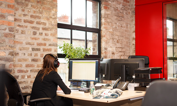 A woman wearing headphones sits in front of a computer in an office space with large windows and exposed brick