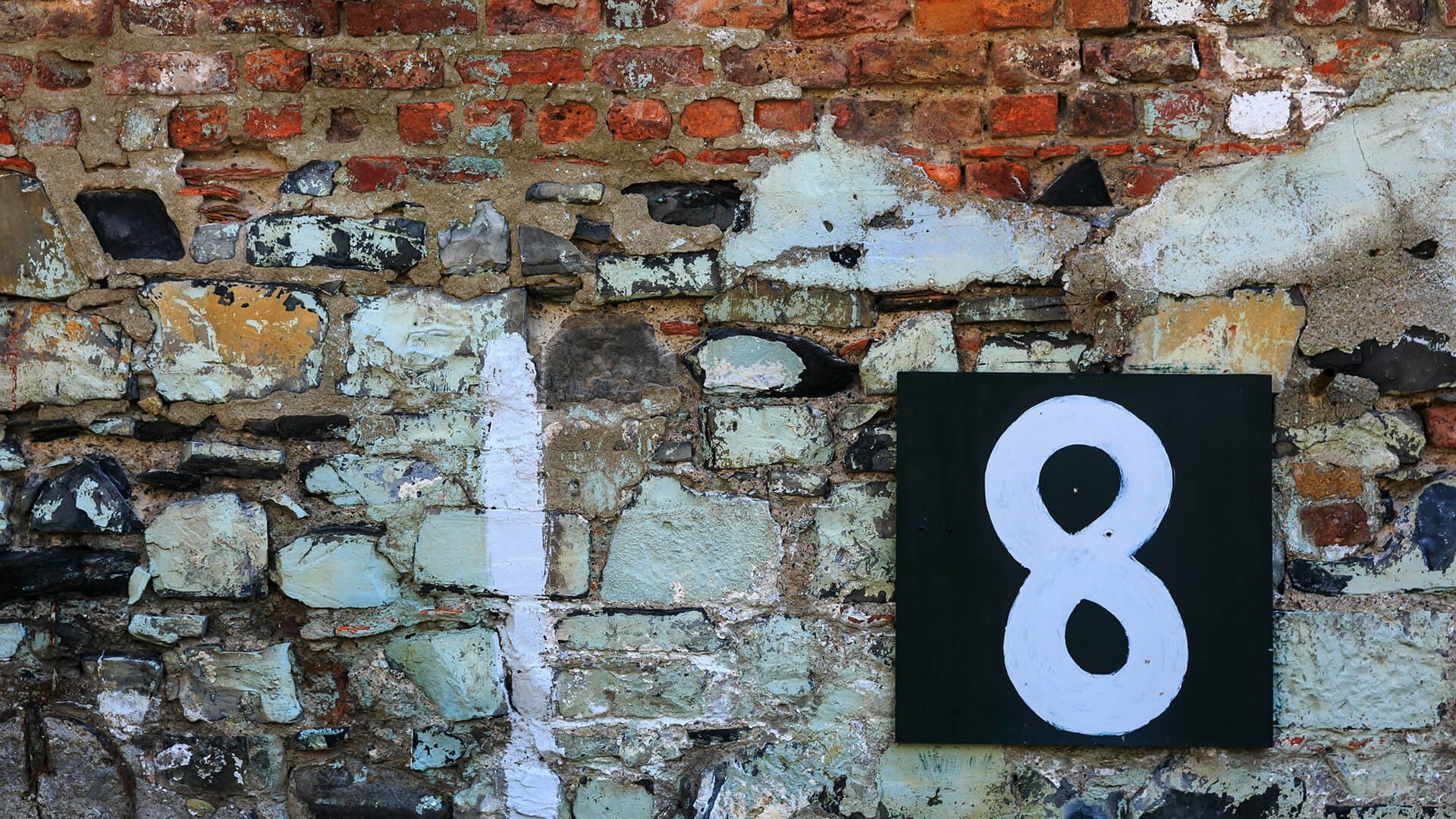 A close-up detail of a colourful, patchwork stone wall. A black sign is mounted on the wall with a number '8' painted on it in white.