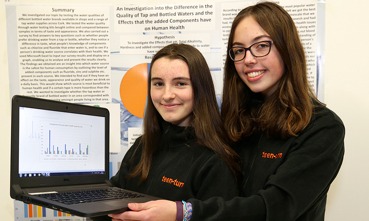 Two smiling teenage girls wearing tops with orange 'Teen Turn' logos hold up a laptop