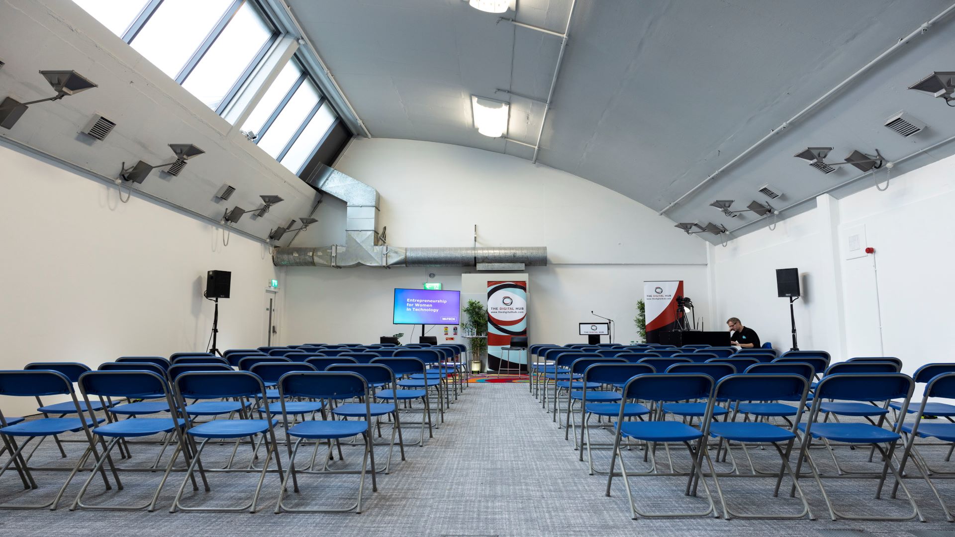 Rows of blue chairs in a meeting room with 'Digital Hub' pullups at the top of the room. There is also a wide screen tv, table and lectern visible. Carpet is grey and bright light comes in from the roof.